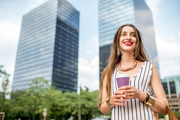 Jeune femme d'affaires souriante prenant une pause-café debout à l'extérieur sur le fond des gratte-ciel de la ville de Bruxelles