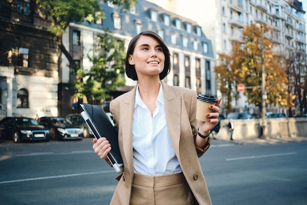 Jeune femme d'affaires souriante et séduisante avec ordinateur portable et café pour aller joyeusement en regardant loin dans la rue de la ville