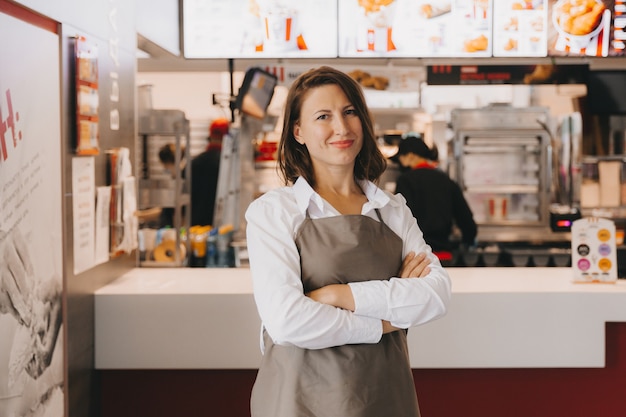 Une jeune femme d'affaires souriante en tablier se tient dans un café devant une vitrine, les bras croisés, regardant la caméra.