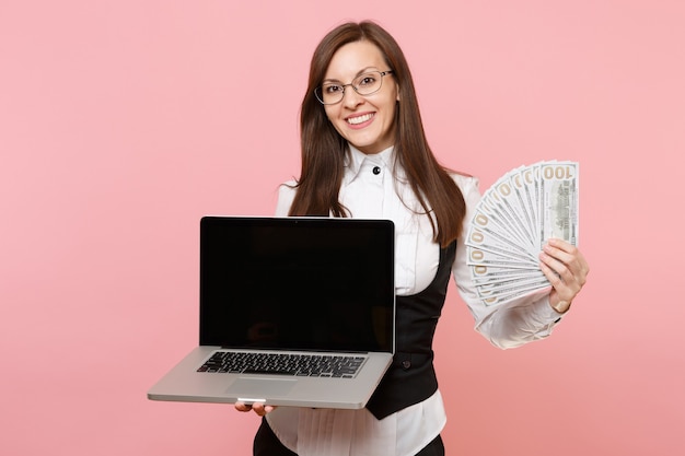 Jeune femme d'affaires souriante tenant un paquet de dollars, de l'argent en espèces et un ordinateur portable avec un écran vide isolé sur fond rose. Dame patronne. Réalisation carrière richesse. Espace de copie.