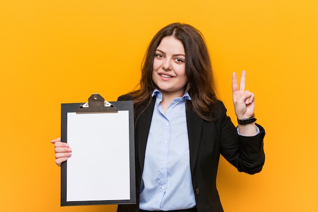 Jeune femme d'affaires de taille plus curvy tenant un presse-papiers montrant le signe de la victoire et souriant largement.