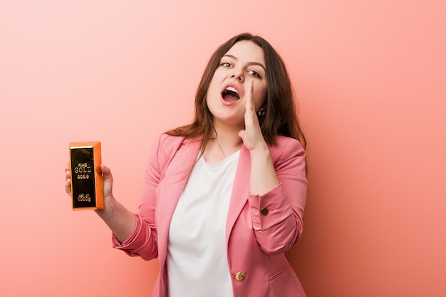 Jeune femme d'affaires de taille plus sinueuse tenant un lingot d'or criant excité à l'avant.