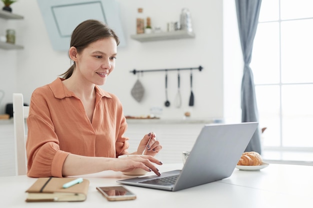 Jeune femme d'affaires tapant sur un ordinateur portable à la table, elle travaille en ligne dans la cuisine