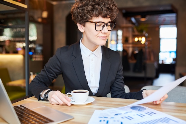 Jeune femme d'affaires avec une tasse de café assis par table au café, la lecture du contrat et la préparation de rendez-vous avec le client