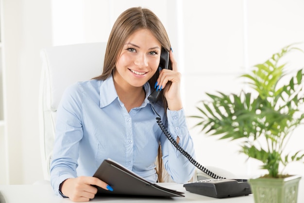 Photo jeune femme d'affaires téléphonant au bureau, assise à un bureau avec une main tenant le téléphone et dans un autre dossier.