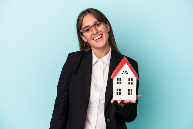 Jeune femme d'affaires tenant une maison de jouet isolée sur fond bleu heureuse, souriante et joyeuse.