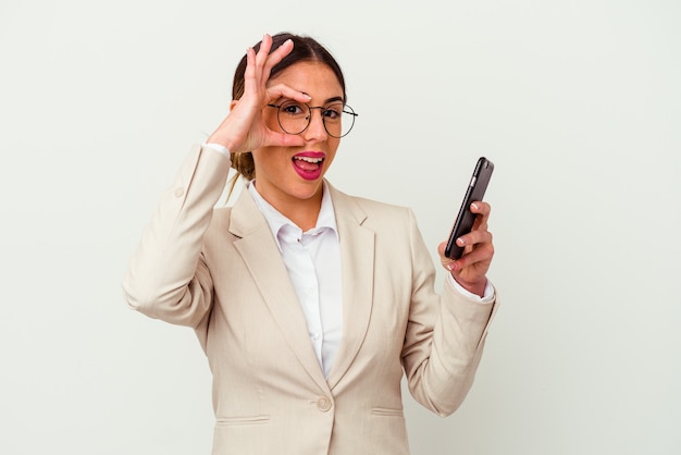 Jeune femme d'affaires tenant un téléphone portable isolé sur blanc excité en gardant un geste ok sur les yeux.