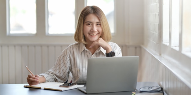 Jeune femme d'affaires travaillant sur son projet avec un ordinateur portable et souriant à la caméra