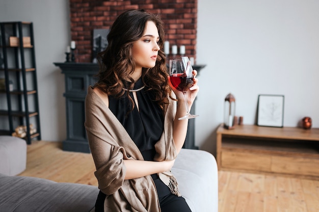 Jeune femme d'affaires travaille à la maison. Un modèle calme et réfléchi tient un verre de vin rouge et attend avec impatience. Portez une robe foncée et un pull marron. Seul dans la chambre.