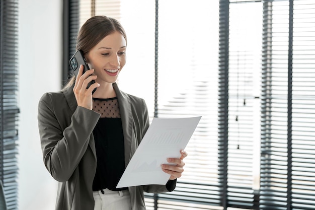 Jeune femme d'affaires utilisant un téléphone portable et regardant un document dans sa main debout dans un bureau moderne