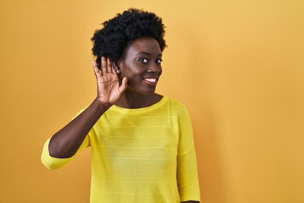 Jeune femme africaine debout au-dessus d'un studio jaune souriant avec la main sur l'oreille écoutant une rumeur ou des commérages. notion de surdité.