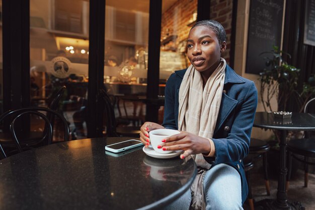 Une jeune femme africaine élégante dans un café en train de boire du café.