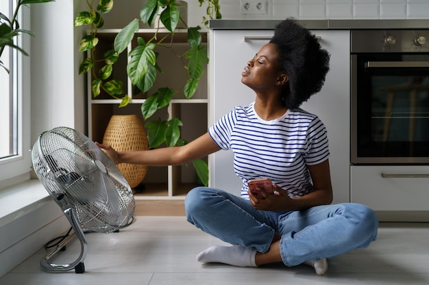 Une jeune femme africaine est assise sur le sol avec un téléphone et capte les courants d'air émanant d'un ventilateur électrique