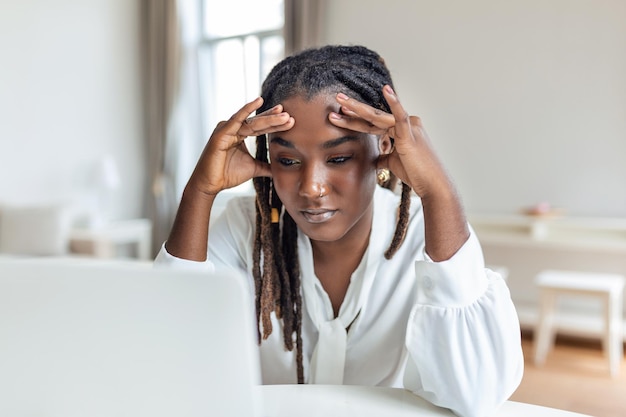 Jeune femme africaine frustrée travaillant au bureau devant un ordinateur portable souffrant de maux de tête quotidiens chroniques