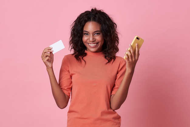 jeune femme africaine montrant une carte de crédit et un téléphone portable sur fond rose