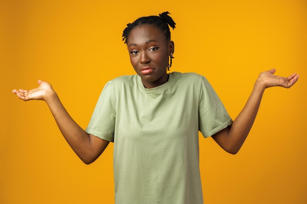 Jeune femme africaine perplexe et désemparée avec les bras en studio jaune