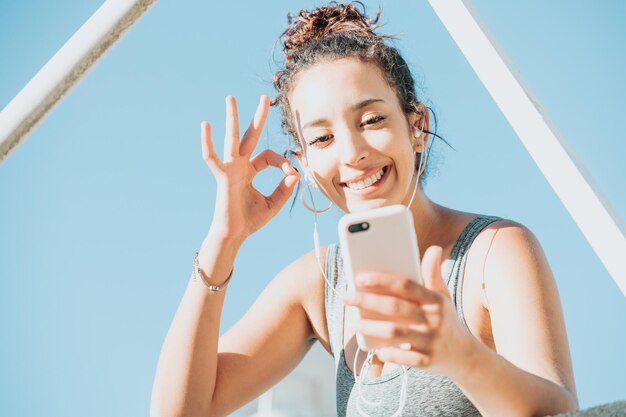 Jeune femme africaine poc assise sur un rebord heureuse après avoir fait beaucoup d'exercice et écouté de la musique, pose cool, avec espace de copie, en leggins de sport et haut. Fille de remise en forme aux cheveux bouclés, signe ok