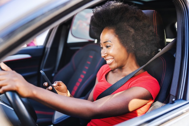 Jeune femme africaine séduisante au volant de sa voiture et regardant un téléphone intelligent.