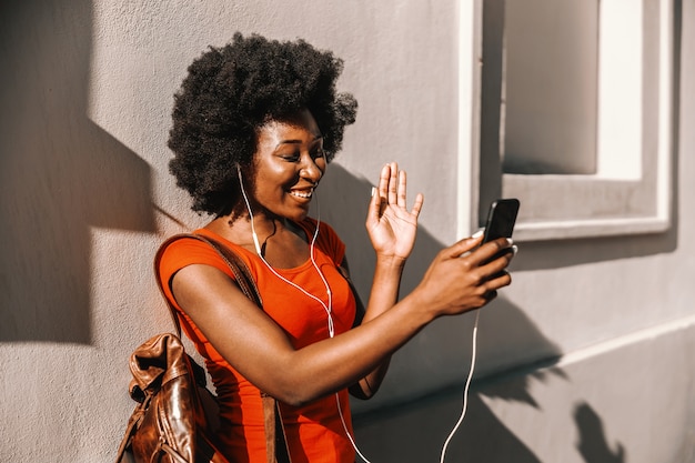 Photo jeune femme africaine séduisante debout à l'extérieur, ayant un appel vidéo sur téléphone intelligent et agitant