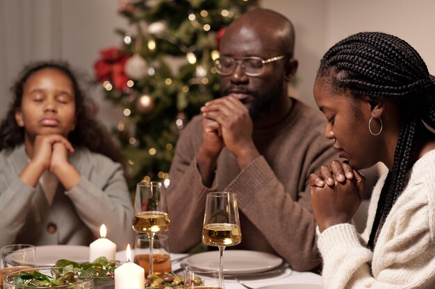 Jeune femme africaine, son mari et leur fille mignonne priant avant le dîner avec leurs mains réunies par le menton