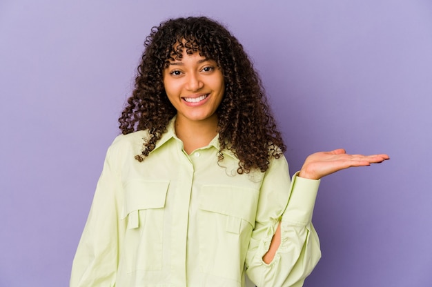 Jeune femme afro-américaine afro-américaine isolée montrant un espace de copie sur une paume et tenant une autre main sur la taille.