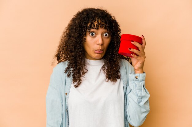 Jeune femme afro-américaine afro-américaine tenant un cadeau de la Saint-Valentin hausse les épaules et les yeux ouverts confus.