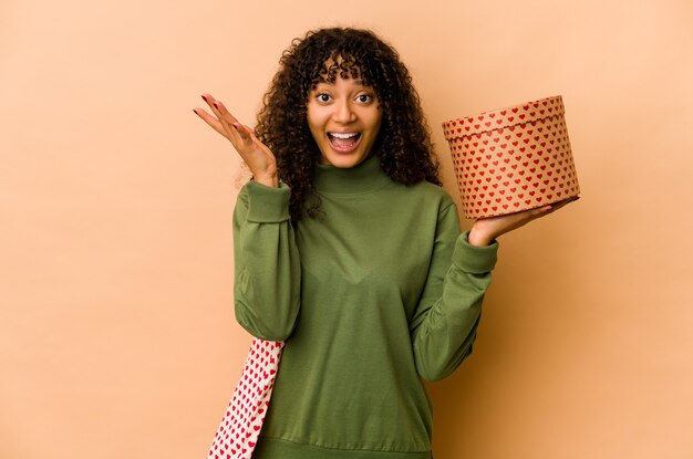 Jeune femme afro-américaine afro-américaine tenant un cadeau de Saint Valentin recevant une agréable surprise, excitée et levant les mains.