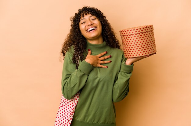 Jeune femme afro-américaine afro-américaine tenant un cadeau de la Saint-Valentin rit fort en gardant la main sur la poitrine.