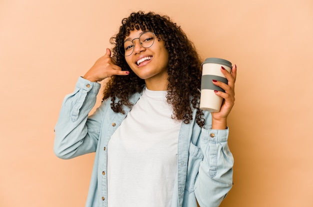 Jeune femme afro-américaine afro-américaine tenant un café à emporter montrant un geste d'appel de téléphone mobile avec les doigts.