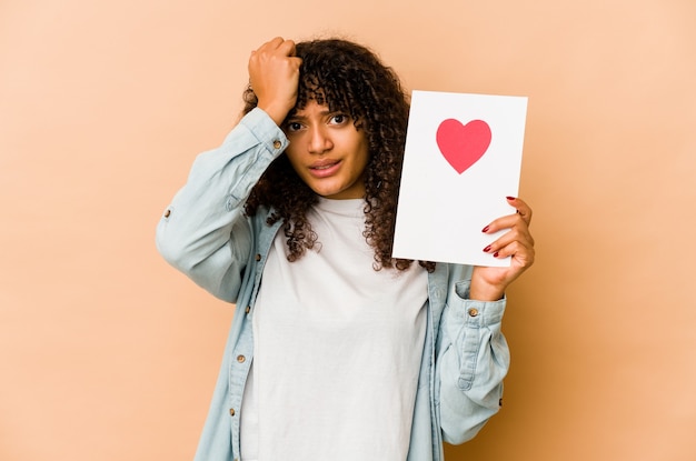 Jeune femme afro-américaine afro-américaine tenant une carte de Saint Valentin choquée, elle s'est souvenue d'une réunion importante.