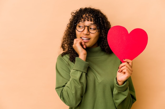 Jeune femme afro-américaine afro-américaine tenant un coeur de la Saint-Valentin détendu penser à quelque chose en regardant un espace de copie.