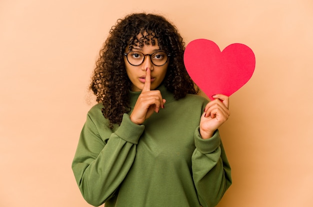 Jeune femme afro-américaine afro-américaine tenant un coeur Saint Valentin gardant un secret ou demandant le silence.