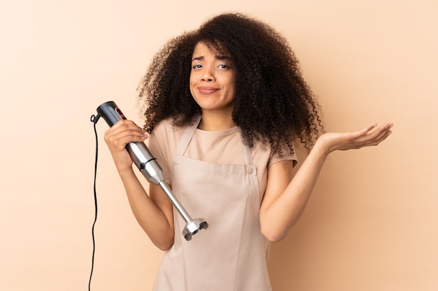 Jeune femme afro-américaine à l'aide d'un mixeur à main avec l'expression du visage confus