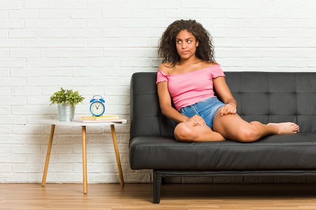 Jeune femme afro-américaine assise sur le canapé, confuse, se sent douteuse et incertaine.