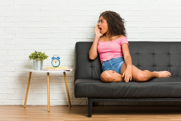 Jeune femme afro-américaine assise sur le canapé en criant et en tenant la paume près de la bouche ouverte.