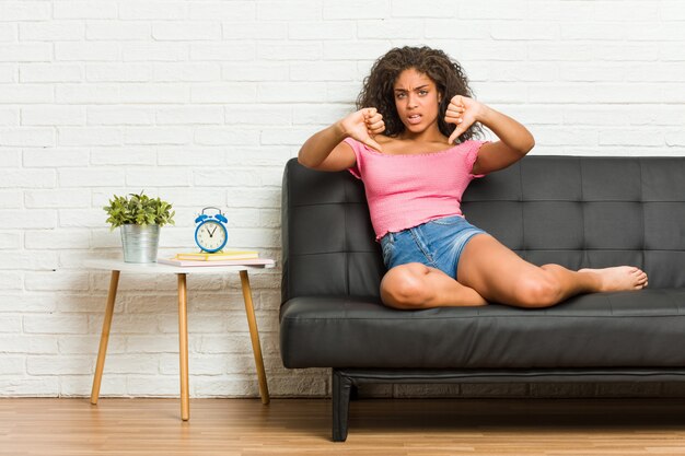 Jeune femme afro-américaine assise sur le canapé montrant le pouce vers le bas et exprimant l'aversion.