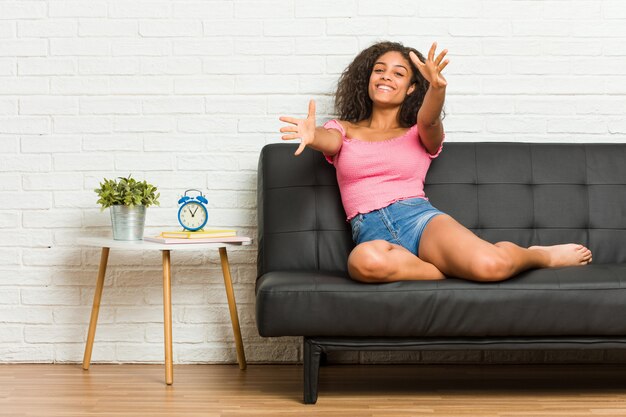 Jeune femme afro-américaine assise sur le canapé se sent confiant en donnant un câlin à la caméra.