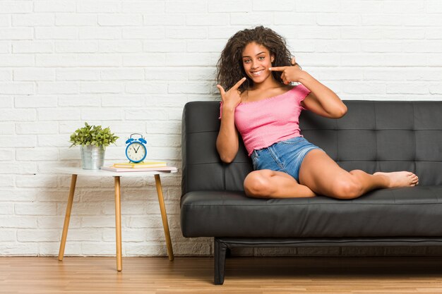 Jeune femme afro-américaine assise sur le canapé sourit, pointer du doigt la bouche.