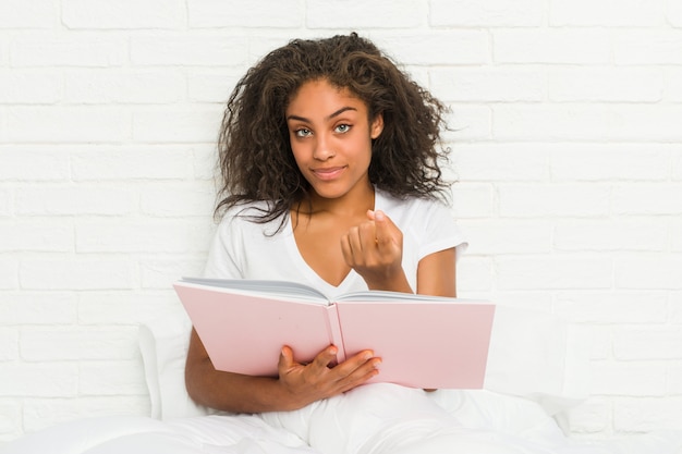 Jeune femme afro-américaine assise sur le lit étudie en pointant du doigt vers vous comme si vous vous y rapprochez