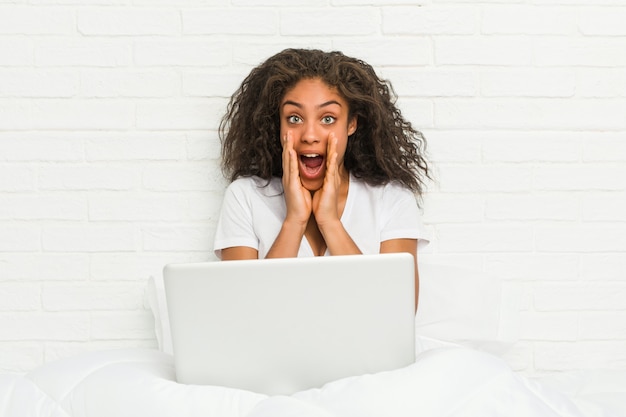 Jeune femme afro-américaine assise sur le lit avec un ordinateur portable criant excité à l'avant.