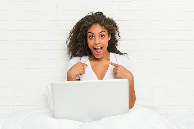 Jeune femme afro-américaine assise sur le lit avec un ordinateur portable surpris en se montrant, souriant largement.