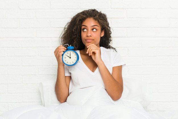 Jeune femme afro-américaine assise sur le lit tenant un réveil à la recherche de côté avec une expression douteuse et sceptique.
