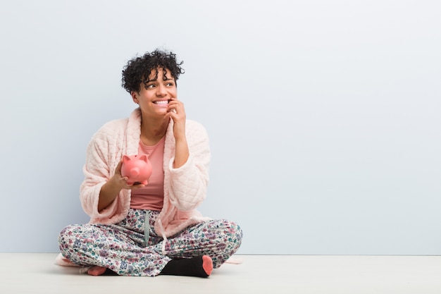 Jeune femme afro-américaine assise avec une tirelire se ronger les ongles, nerveuse et très anxieuse.