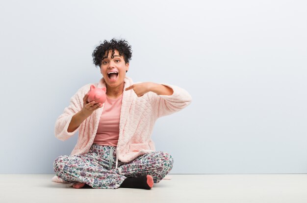 Jeune femme afro-américaine assise avec une tirelire surpris pointant vers lui-même, souriant largement.