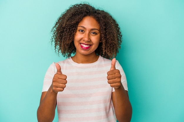 Jeune femme afro-américaine aux cheveux bouclés isolé sur fond bleu souriant et levant le pouce vers le haut