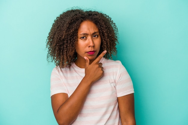 Jeune femme afro-américaine aux cheveux bouclés isolée sur fond bleu contemplant, planifiant une stratégie, réfléchissant à la manière d'une entreprise.