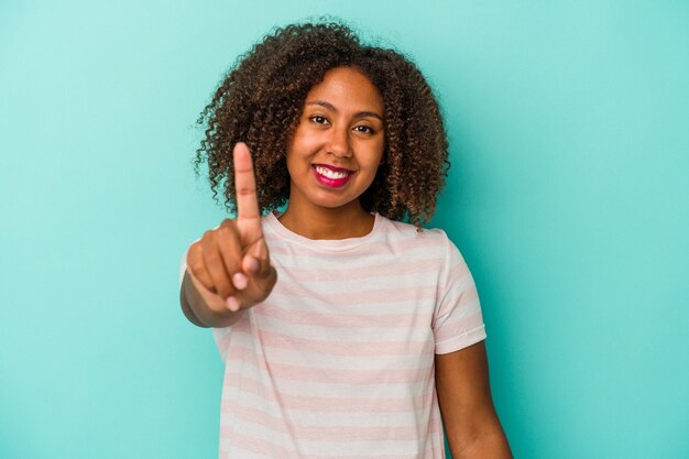 Photo jeune femme afro-américaine aux cheveux bouclés isolée sur fond bleu montrant le numéro un avec le doigt.