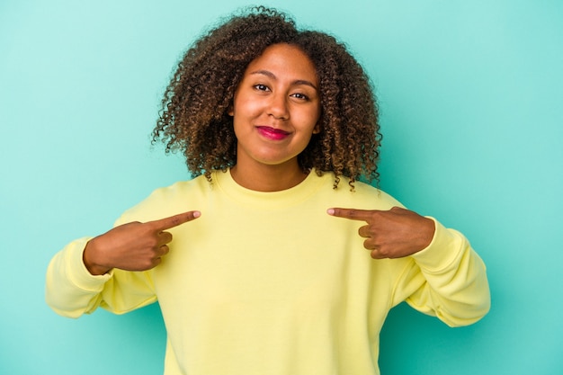 Jeune femme afro-américaine aux cheveux bouclés isolée sur fond bleu personne pointant à la main vers un espace de copie de chemise, fière et confiante