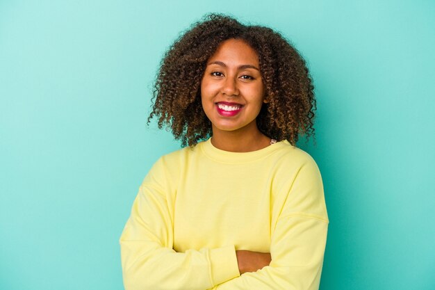 Jeune femme afro-américaine aux cheveux bouclés isolée sur fond bleu qui se sent confiante, croisant les bras avec détermination.