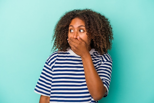 Jeune femme afro-américaine aux cheveux bouclés isolée sur fond bleu réfléchie à la recherche d'un espace de copie couvrant la bouche avec la main.